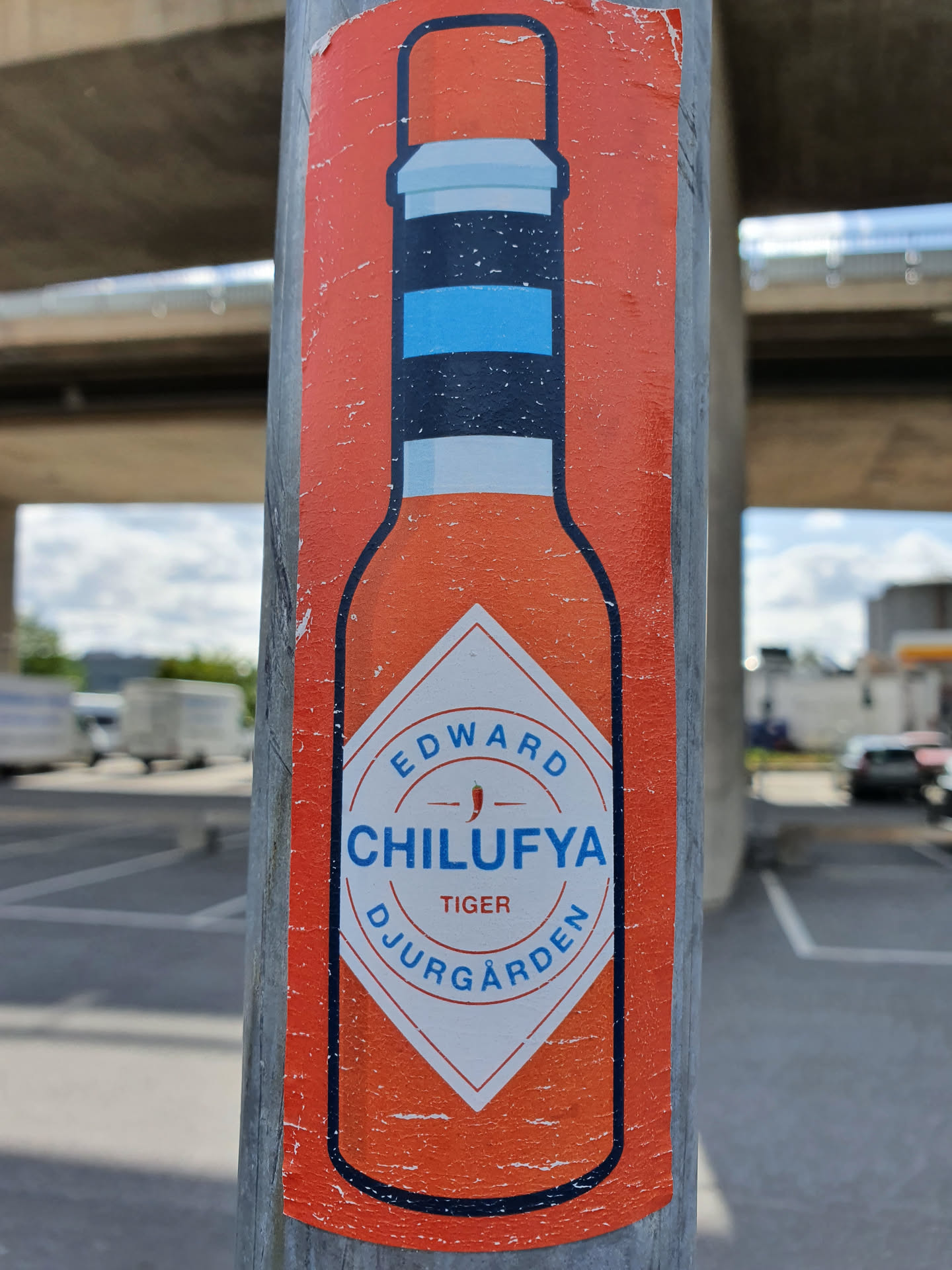 A sticker featuring a hot sauce bottle design with the text "Edward Chilufya", "Tiger", and "Djurgården". The sticker is affixed to a metal pole.
