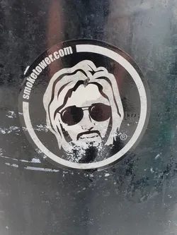 Black and white sticker featuring a stylized illustration of a person with long hair and sunglasses. The text "SmokeTower.com" is visible on the sticker.