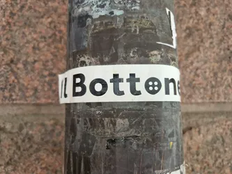 A white sticker with the word "Bottonet" in bold black text and a small button icon. The sticker is attached to a dark-colored pole outdoors against a stone wall. 
