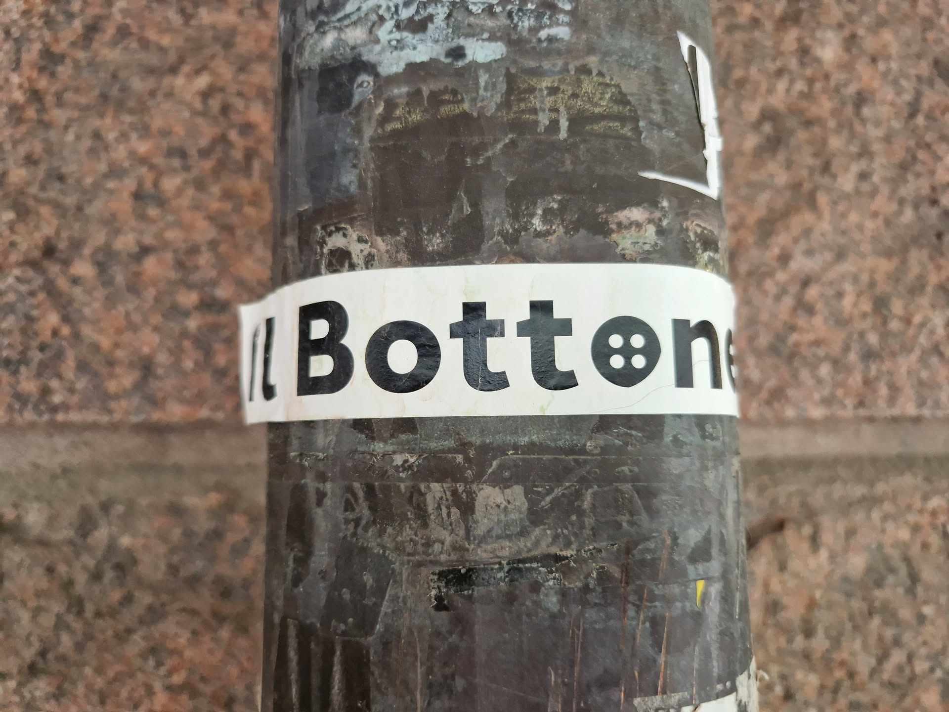 A white sticker with the word "Bottonet" in bold black text and a small button icon. The sticker is attached to a dark-colored pole outdoors against a stone wall. 
