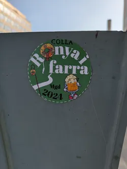 A round sticker with green background features a wreath, a walking stick, and a beer mug character. It mentions Colla Ronyai Farra MGD 2024.