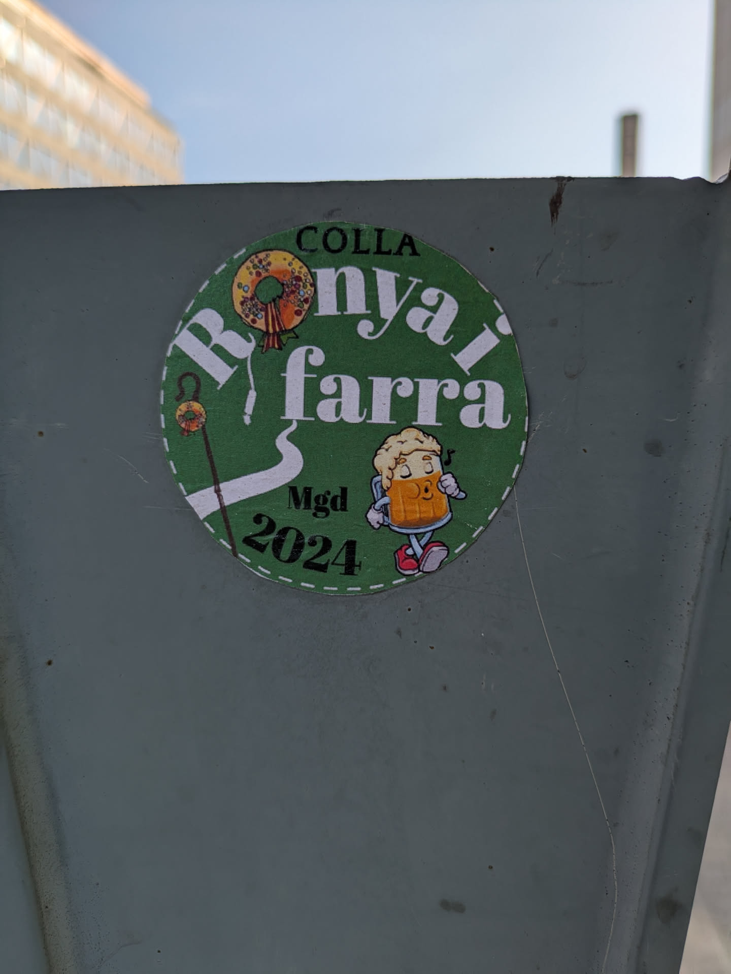 A round sticker with green background features a wreath, a walking stick, and a beer mug character. It mentions Colla Ronyai Farra MGD 2024.