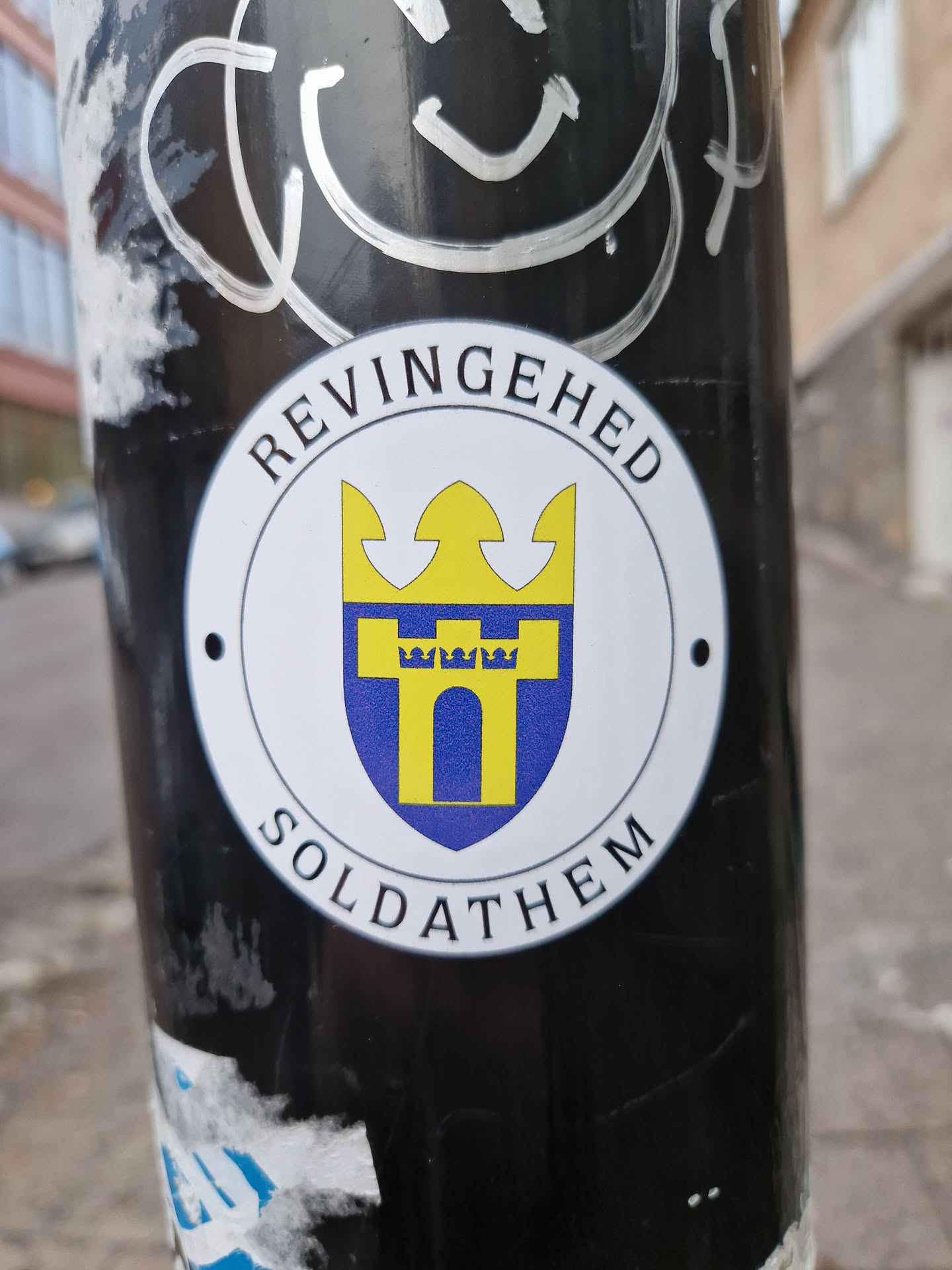Close-up of a circular sticker displaying the coat of arms of Soldathem, a locality in Revingehed, Sweden. The emblem showcases a yellow and blue shield with a castle-like structure and a crown. The text 'REVINGEHED' and 'SOLDATHEM' surrounds the emblem.