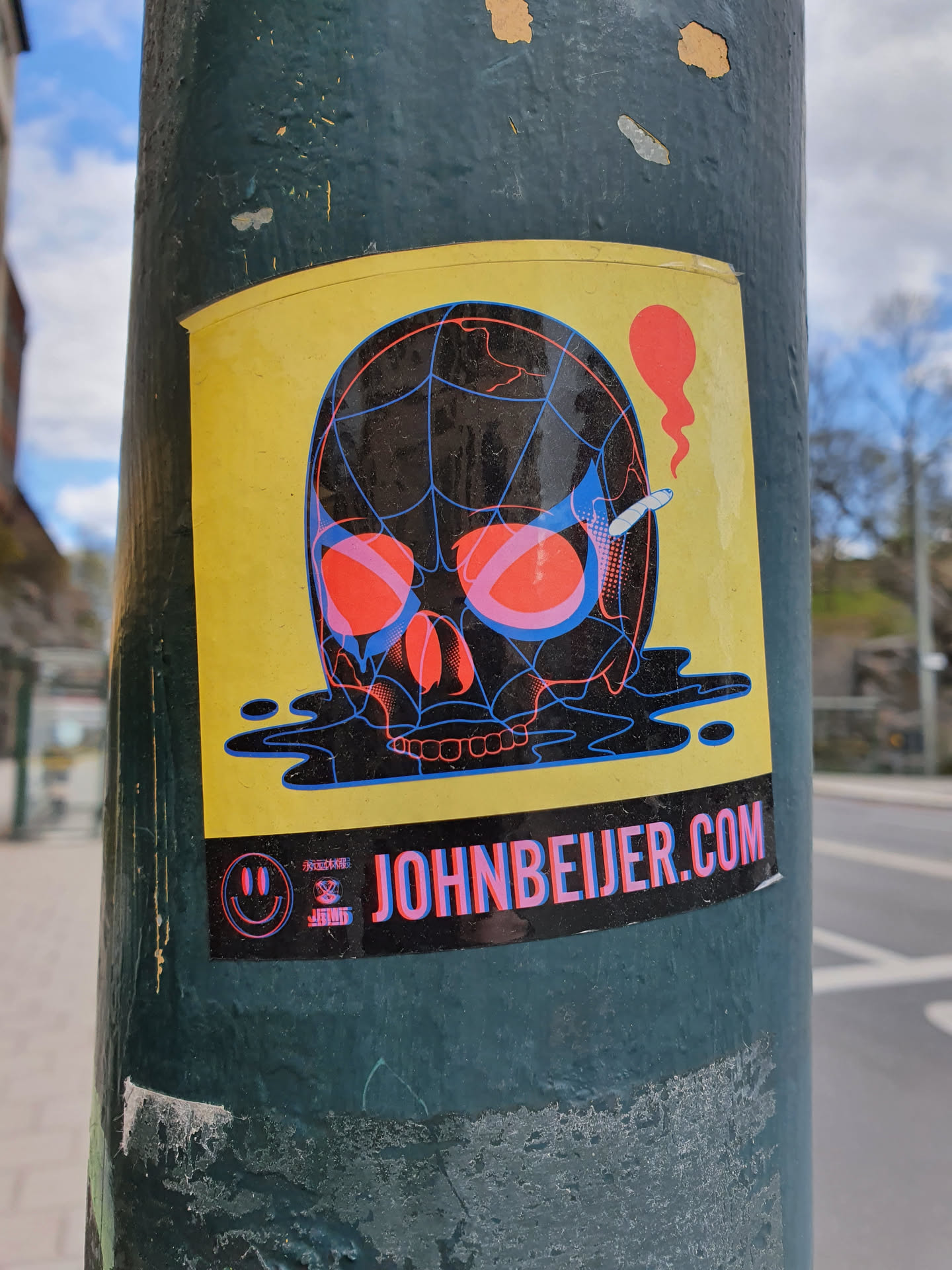 Sticker featuring a skull design with a spiderweb pattern, red eyes, and a cigarette. The skull is depicted melting, and the background is yellow. The sticker includes the website JOHNBEIJER.COM and a smiley face logo. 