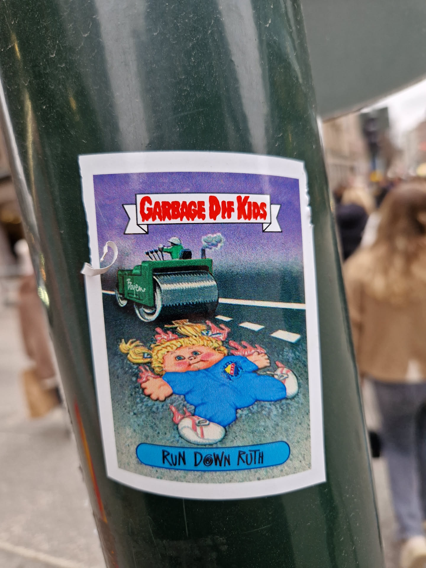 A sticker featuring a Garbage Pail Kids card.  The card depicts a Cabbage Patch Kid being run over by a steamroller. The text on the card says "Garbage Pail Kids" and "Run Down Ruth".