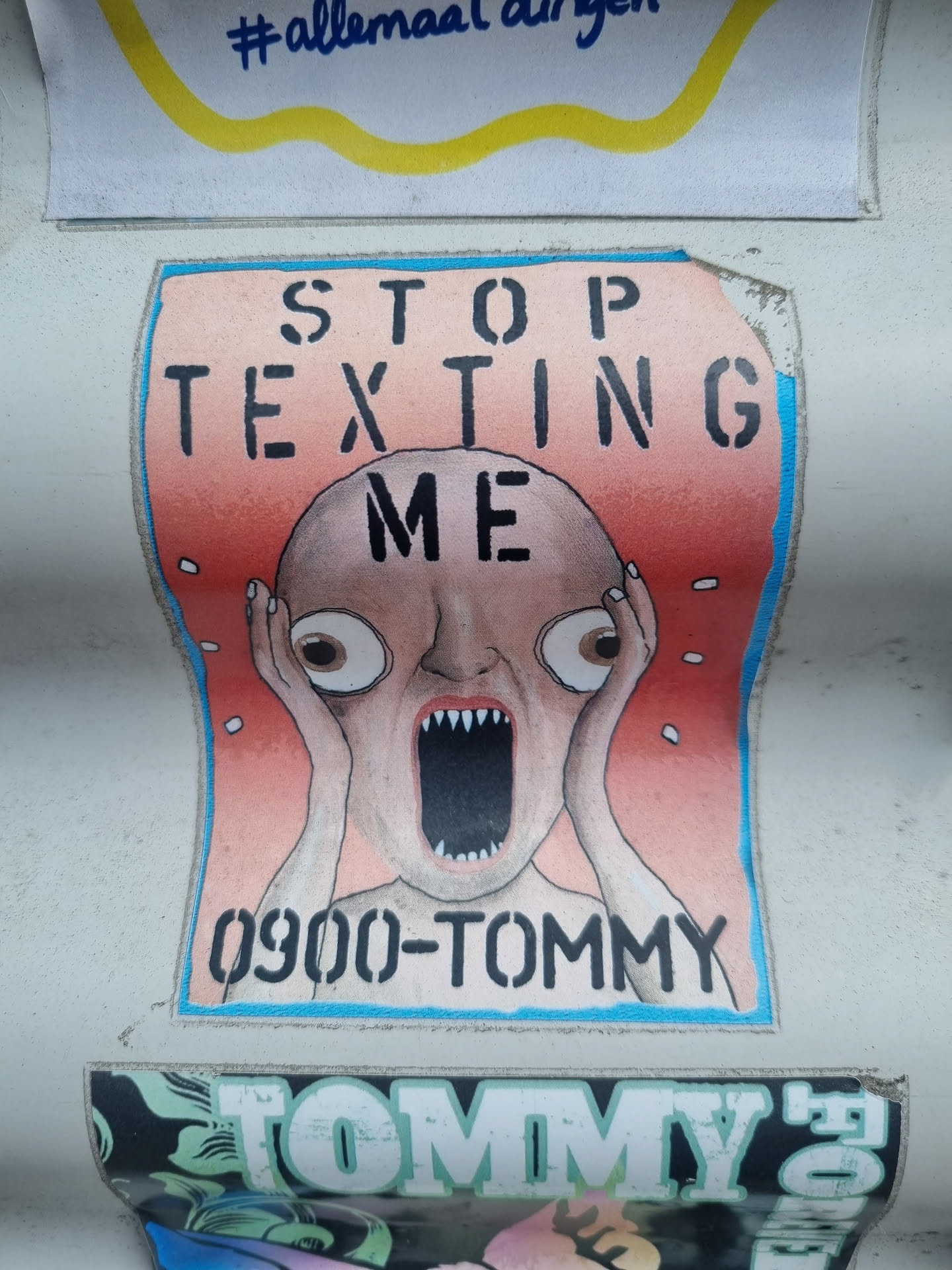 A sticker featuring a screaming bald head with the text 'STOP TEXTING ME' and a phone number '0900-TOMMY'. The art style is cartoonish and slightly disturbing. The sticker is stuck on a wall or surface among other stickers.