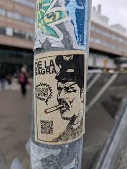A sticker featuring a black and white illustration of a man smoking a cigarette. The text on the sticker says "De La Sagrada" and "Unfiltered Music of Woe!"  There is a QR code at the bottom.