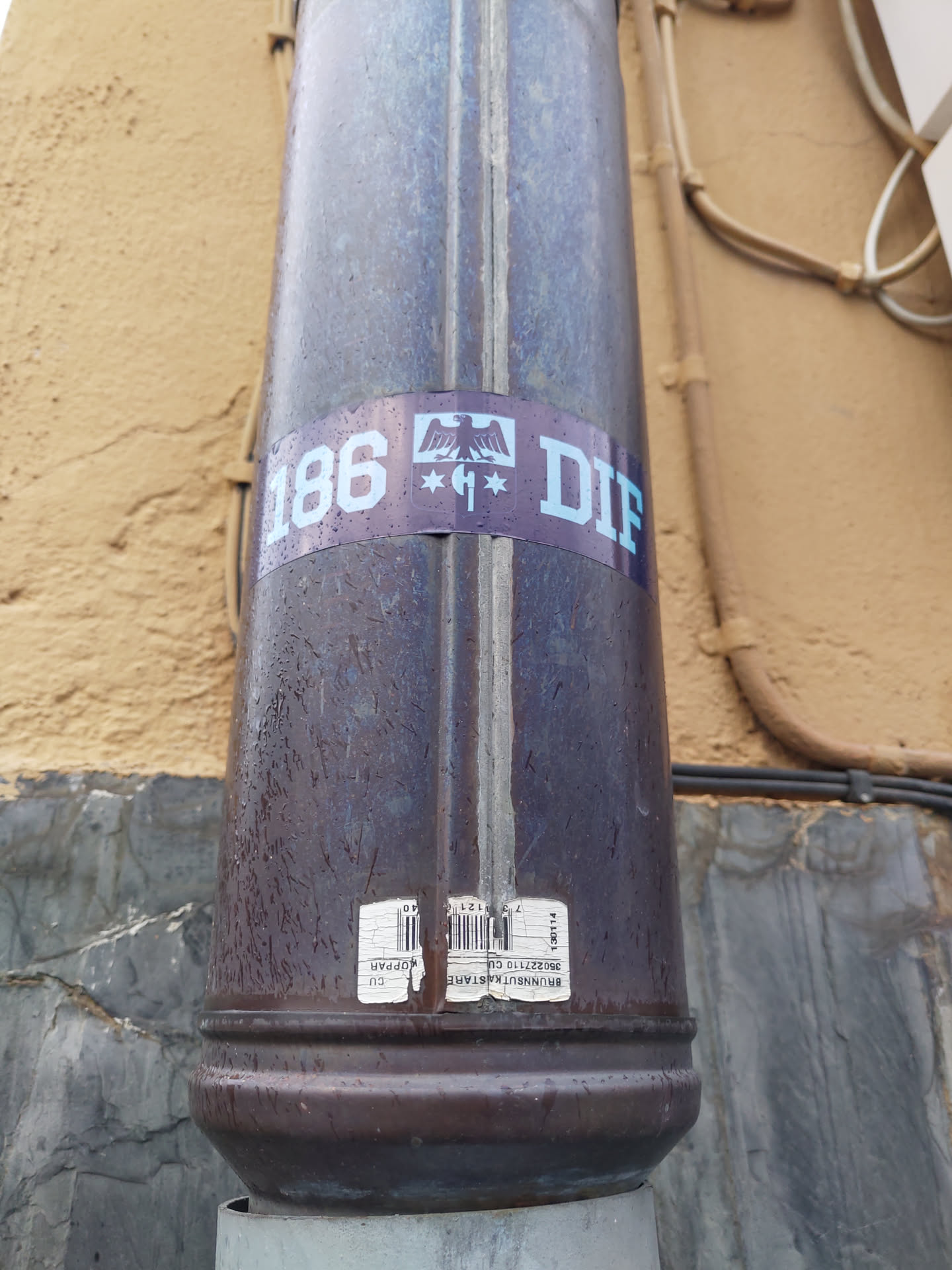 A sticker featuring the number 186, a stylized German eagle, and the letters DIF.  The sticker is affixed to a dark brown metal downspout. The style is minimalistic and possibly related to a specific group or movement.