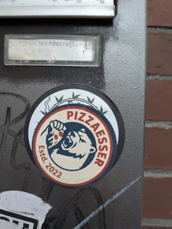 A circular logo sticker placed over another round sticker on a gray metal surface. The sticker has an off-white background framed by navy blue and red rings. At the center is a navy blue silhouette illustration of a roaring bear head preparing to eat a dripping slice of pizza adorned with red pepperoni. Arched text in red capital letters reads 'PIZZAESSER' along the top right, while 'Estd. 2022' is written along the lower left edge.