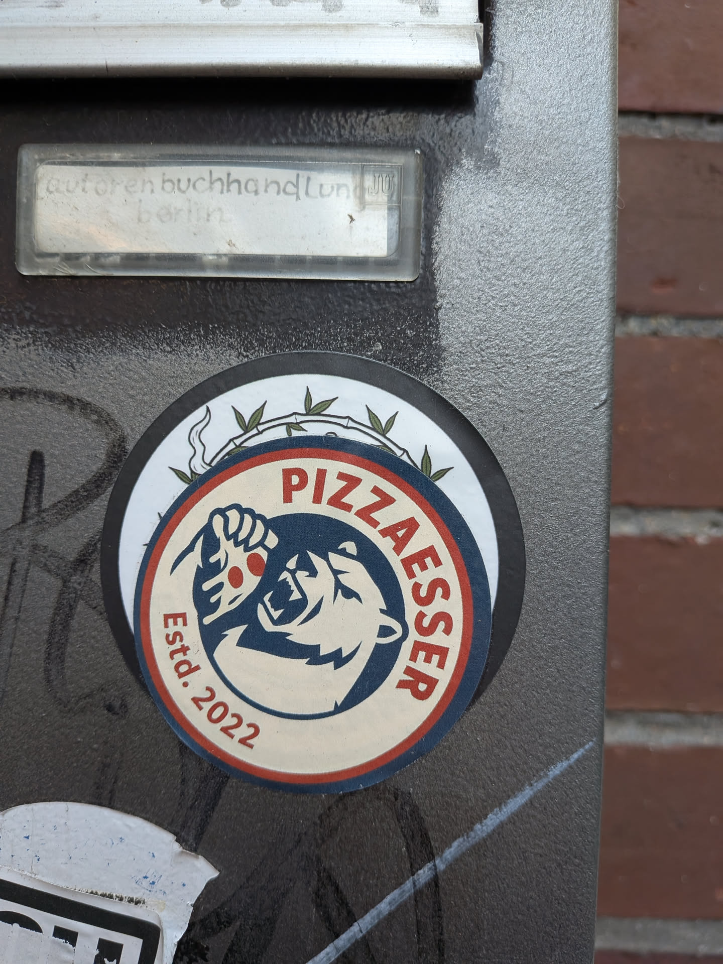 A circular logo sticker placed over another round sticker on a gray metal surface. The sticker has an off-white background framed by navy blue and red rings. At the center is a navy blue silhouette illustration of a roaring bear head preparing to eat a dripping slice of pizza adorned with red pepperoni. Arched text in red capital letters reads 'PIZZAESSER' along the top right, while 'Estd. 2022' is written along the lower left edge.
