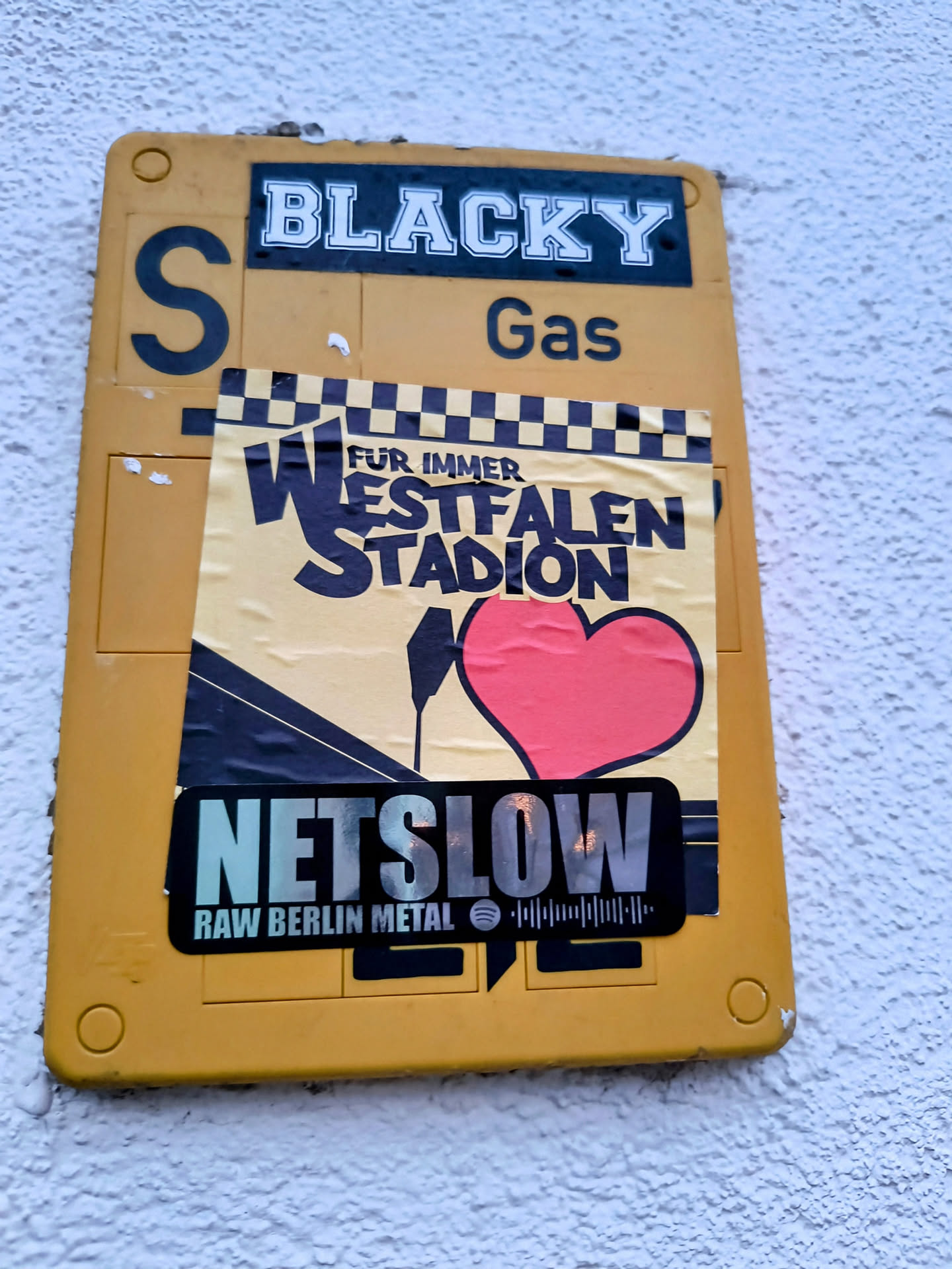 Sticker on a yellow box. The sticker has the band name NETSLOW and the text RAW BERLIN METAL. There is also a Spotify code.  Above it is another sticker with Westfalenstadion and a heart.
