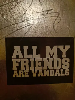 A black sticker with white text that says "All my friends are vandals". It's been placed on a wall with other markings.