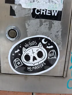 The sticker is oval-shaped with a black background and a distinct white border. Its central motif is a stylized white cat skull with large, empty black eye sockets, a small nose, and jagged teeth. A cross symbol is on its forehead. Above the skull, the text 'No GODS' is written in white, and below it, 'NO MASTERS' is written, also in white. The background features a night sky theme with a crescent moon and stars on the right, and a bat flying on both the left and right sides. Two gravestones are depicted at the bottom: one on the left with '666' and a star, and another on the right with 'R.I.P.'. The ground beneath the gravestones is represented by a dotted pattern. The overall aesthetic is black and white, giving it a stark, graphic appearance.