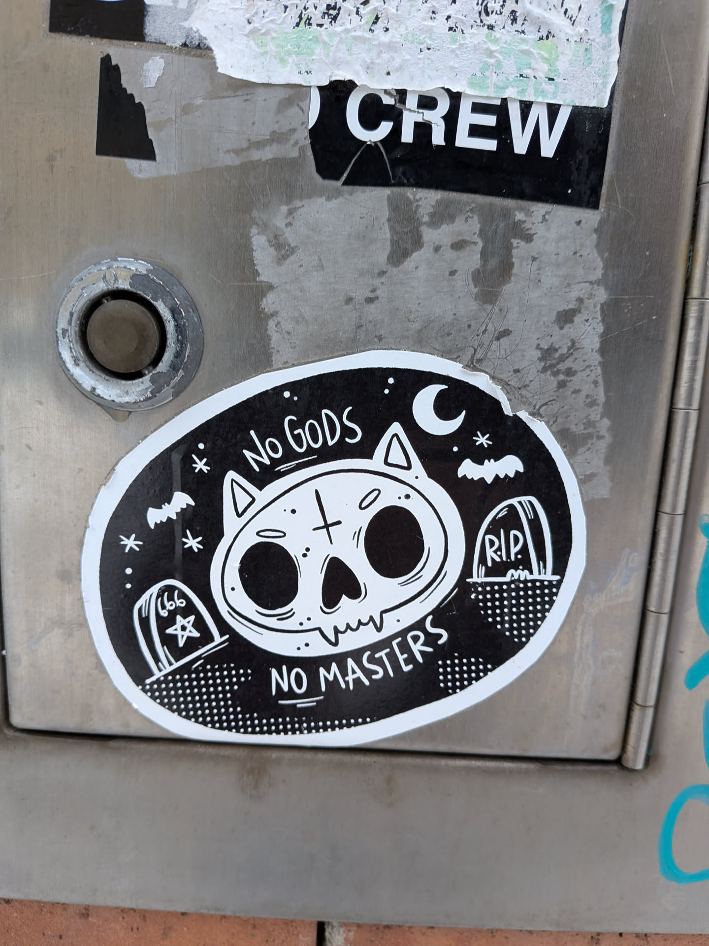 The sticker is oval-shaped with a black background and a distinct white border. Its central motif is a stylized white cat skull with large, empty black eye sockets, a small nose, and jagged teeth. A cross symbol is on its forehead. Above the skull, the text 'No GODS' is written in white, and below it, 'NO MASTERS' is written, also in white. The background features a night sky theme with a crescent moon and stars on the right, and a bat flying on both the left and right sides. Two gravestones are depicted at the bottom: one on the left with '666' and a star, and another on the right with 'R.I.P.'. The ground beneath the gravestones is represented by a dotted pattern. The overall aesthetic is black and white, giving it a stark, graphic appearance.