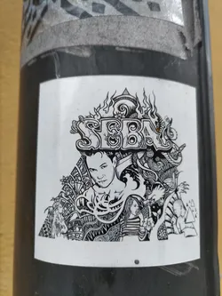 A black and white sticker with the word SEBA prominently displayed in a stylized font. The design is detailed, incorporating various elements such as flames, swirling patterns, and figures.  A central male figure is depicted, surrounded by an array of smaller figures and detailed designs.