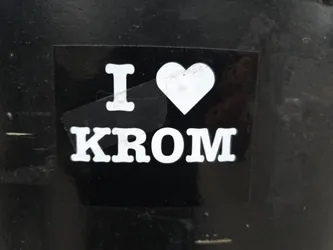A black and white sticker with the text "I \u2764 KROM" on it. The heart is white, and the text is white against a black background. The sticker is attached to a dark surface.