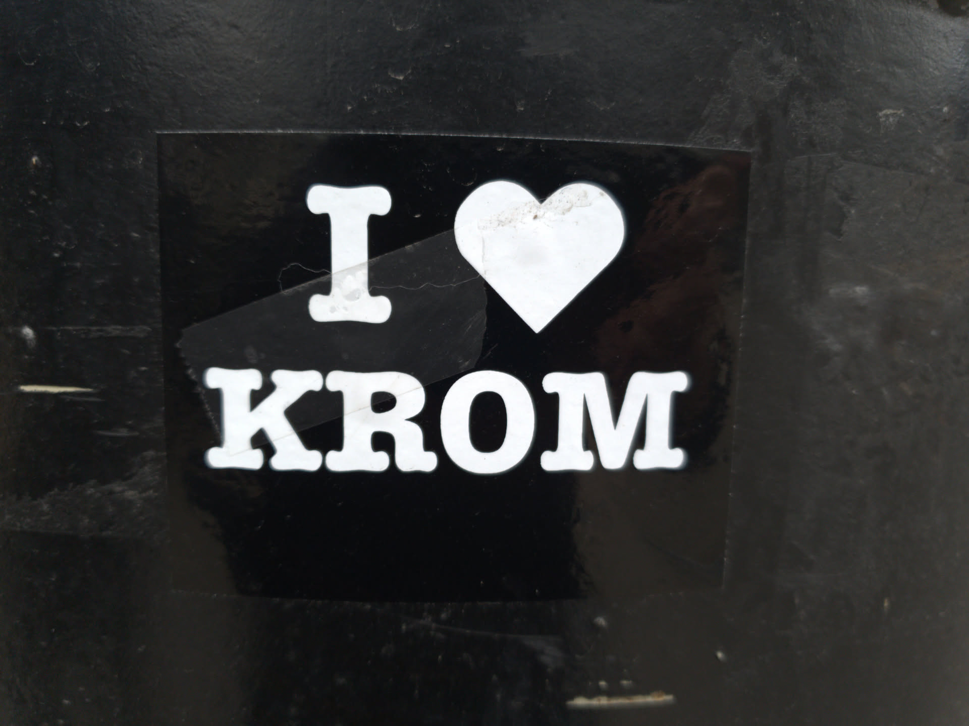 A black and white sticker with the text "I \u2764 KROM" on it. The heart is white, and the text is white against a black background. The sticker is attached to a dark surface.