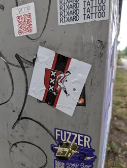The sticker features the crest of AFC Ajax, a Dutch football club. It displays the team's colors: red, black, and white. The crest is a shield with three X's in black on a red background, flanked by vertical black and red stripes. A stylized depiction of a footballer in a red and white striped jersey is shown kicking a soccer ball. The sticker is white with the design elements in red, black, and white, and appears to be slightly damp, with water droplets visible on its surface. It is affixed to a metal post covered in graffiti.
