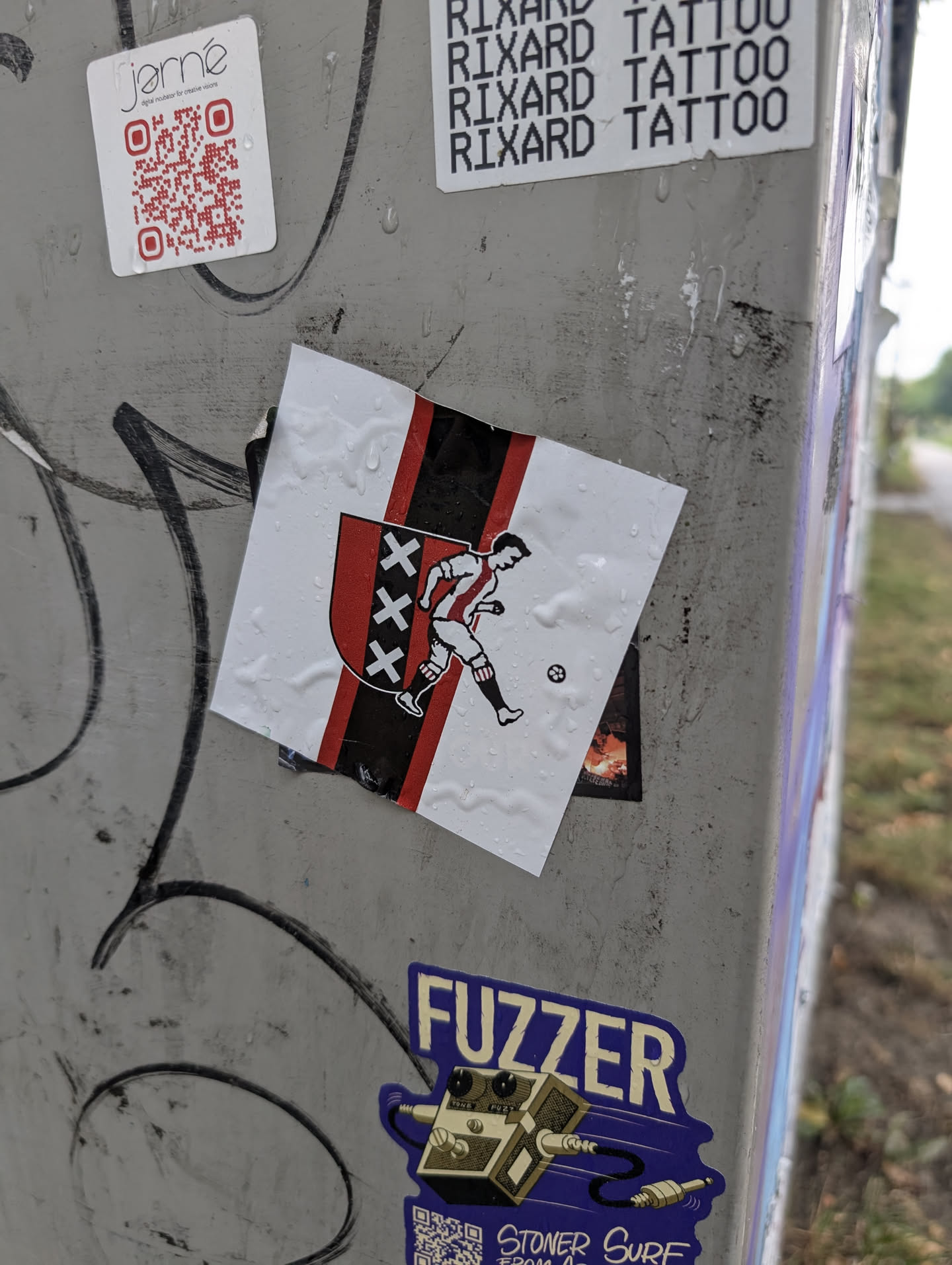 The sticker features the crest of AFC Ajax, a Dutch football club. It displays the team's colors: red, black, and white. The crest is a shield with three X's in black on a red background, flanked by vertical black and red stripes. A stylized depiction of a footballer in a red and white striped jersey is shown kicking a soccer ball. The sticker is white with the design elements in red, black, and white, and appears to be slightly damp, with water droplets visible on its surface. It is affixed to a metal post covered in graffiti.