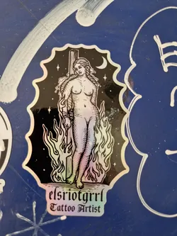 This holographic sticker features a detailed illustration of a woman bound to a stake, surrounded by flames. The background is black with stars and a crescent moon.  The sticker includes the text "elsriotgrrrl Tattoo Artist" at the bottom. The art style is reminiscent of vintage or medieval imagery.