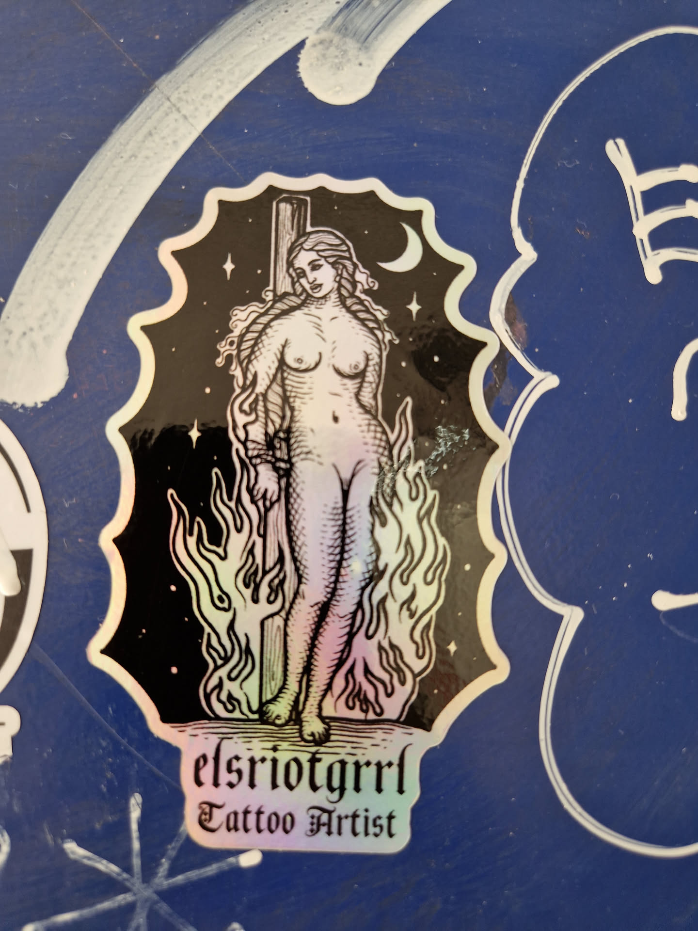 This holographic sticker features a detailed illustration of a woman bound to a stake, surrounded by flames. The background is black with stars and a crescent moon.  The sticker includes the text "elsriotgrrrl Tattoo Artist" at the bottom. The art style is reminiscent of vintage or medieval imagery.