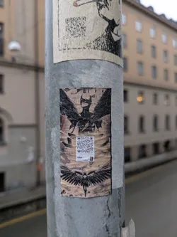 The sticker displays a stylized figure with wings spread above and below. There is a QR code in the center of the figure. The pole appears to be in an urban setting.