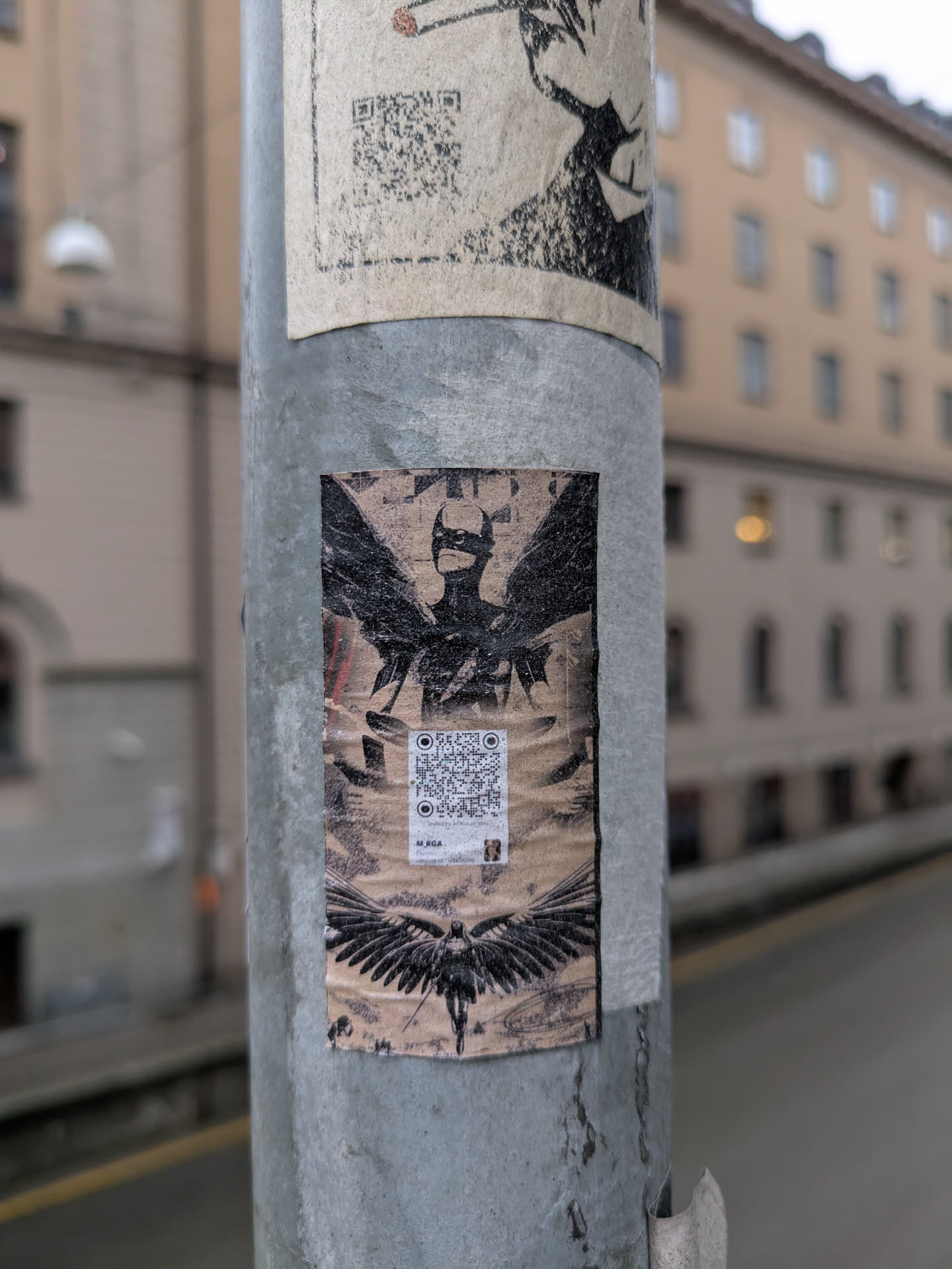The sticker displays a stylized figure with wings spread above and below. There is a QR code in the center of the figure. The pole appears to be in an urban setting.
