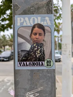 This is a poster attached to a grey metal post. The poster has a pale blue border with rounded corners. At the top, the name 'PAOLA' is printed in large, white, sans-serif letters. Below the name is a portrait of a woman with a serious expression. She is wearing a black top with yellow flower-like pattern. The background of the portrait features what appears to be an interior setting. At the bottom, 'VALENCIA' is printed in large, bold, white letters on a pink background. To the right of 'VALENCIA' is a green circle containing the numbers '20' over '25'.