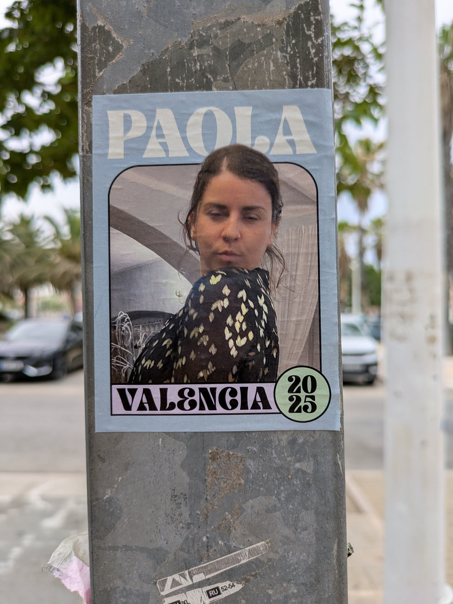 This is a poster attached to a grey metal post. The poster has a pale blue border with rounded corners. At the top, the name 'PAOLA' is printed in large, white, sans-serif letters. Below the name is a portrait of a woman with a serious expression. She is wearing a black top with yellow flower-like pattern. The background of the portrait features what appears to be an interior setting. At the bottom, 'VALENCIA' is printed in large, bold, white letters on a pink background. To the right of 'VALENCIA' is a green circle containing the numbers '20' over '25'.