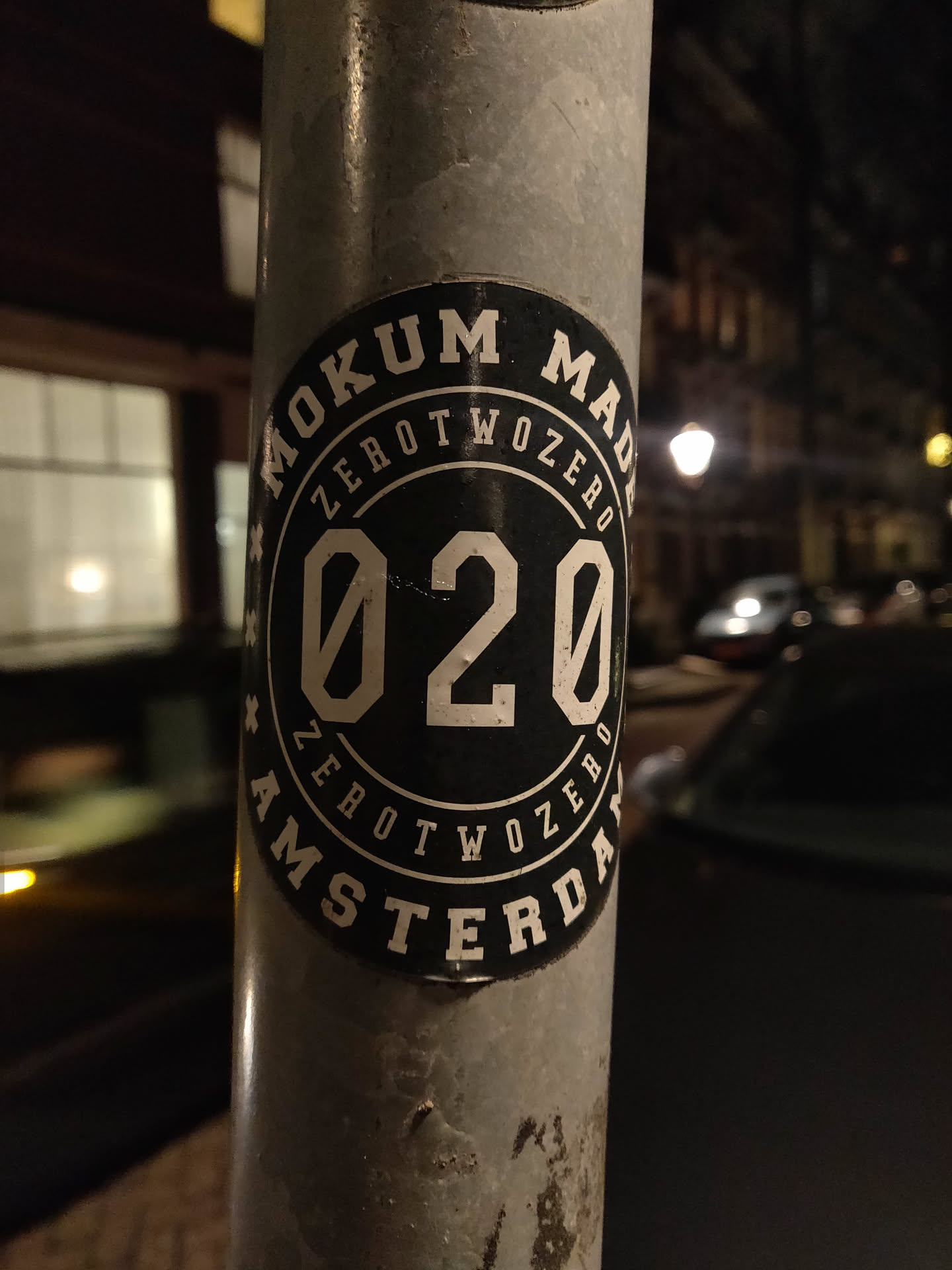 A black and white circular sticker with the text 'Mokum Made' at the top and 'Amsterdam' at the bottom.  In the center is '020' in a bold font.  The sticker is affixed to a metal pole at night.