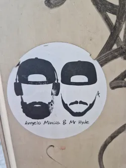 A round sticker featuring black and white minimalist illustrations of two men, possibly Angelo Minoida and Mr Hyde, each wearing a baseball cap. The style is simple and graphic.