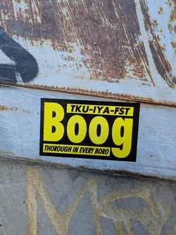 A rectangular black sticker printed with vivid yellow graphic text. Prominently in the center is the name 'Boog' in large, bold sans-serif lettering. Above it is a yellow rectangular box containing the black text 'TKU-IYA-FST'. Beneath it is another yellow rectangular banner reading 'THOROUGH IN EVERY BORO' in black capital letters. The sticker is placed on a weathered, rusty, light-blue painted metal surface.