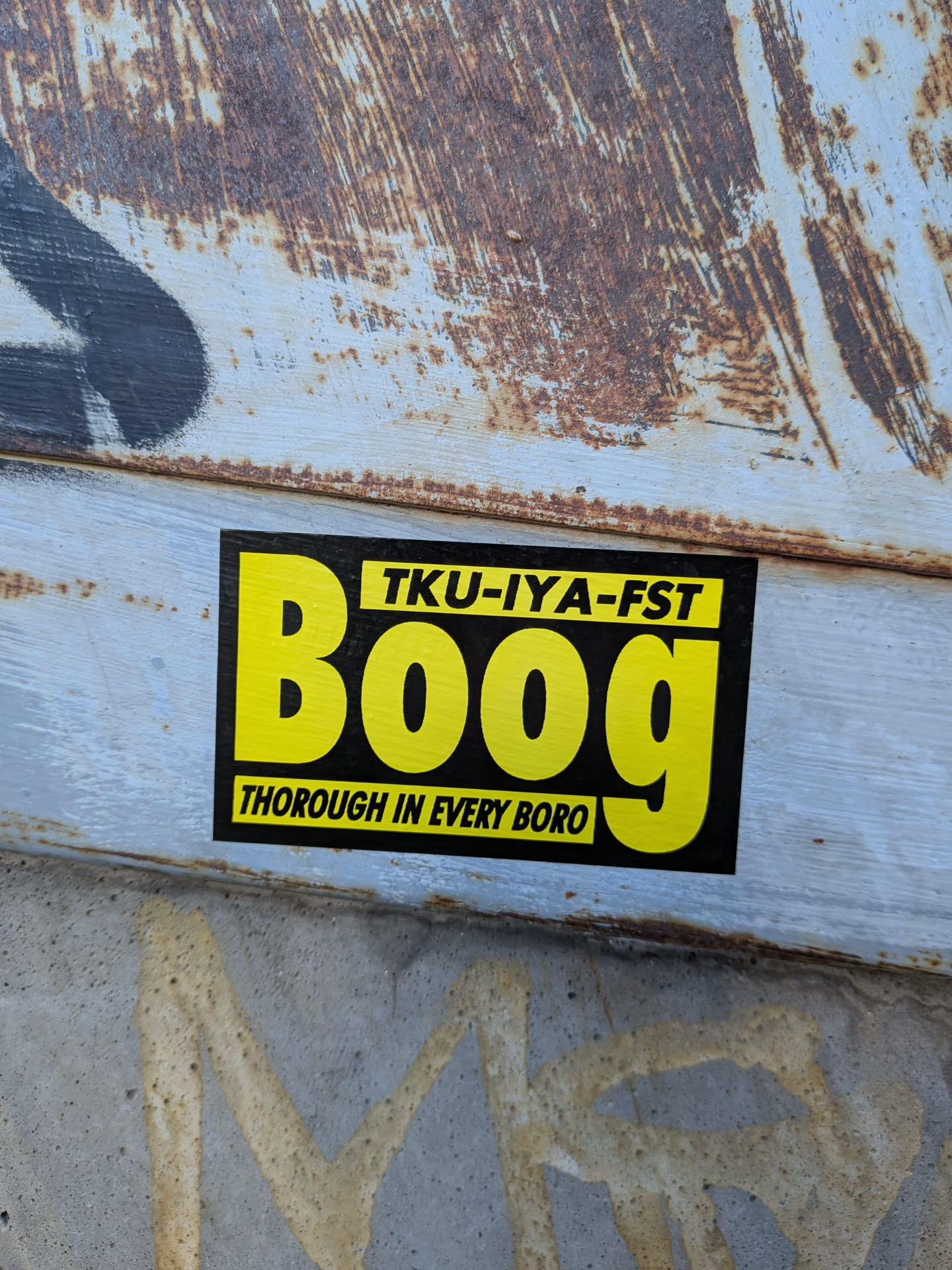 A rectangular black sticker printed with vivid yellow graphic text. Prominently in the center is the name 'Boog' in large, bold sans-serif lettering. Above it is a yellow rectangular box containing the black text 'TKU-IYA-FST'. Beneath it is another yellow rectangular banner reading 'THOROUGH IN EVERY BORO' in black capital letters. The sticker is placed on a weathered, rusty, light-blue painted metal surface.