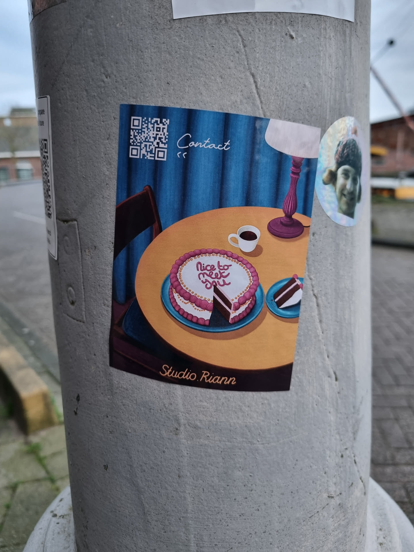 A sticker featuring an illustration of a heart-shaped cake on a table with a cup of coffee, a lamp, and a chair. The text "Nice to meet you" is written on the cake. The sticker has a QR code for contact and is signed "Studio Riann".