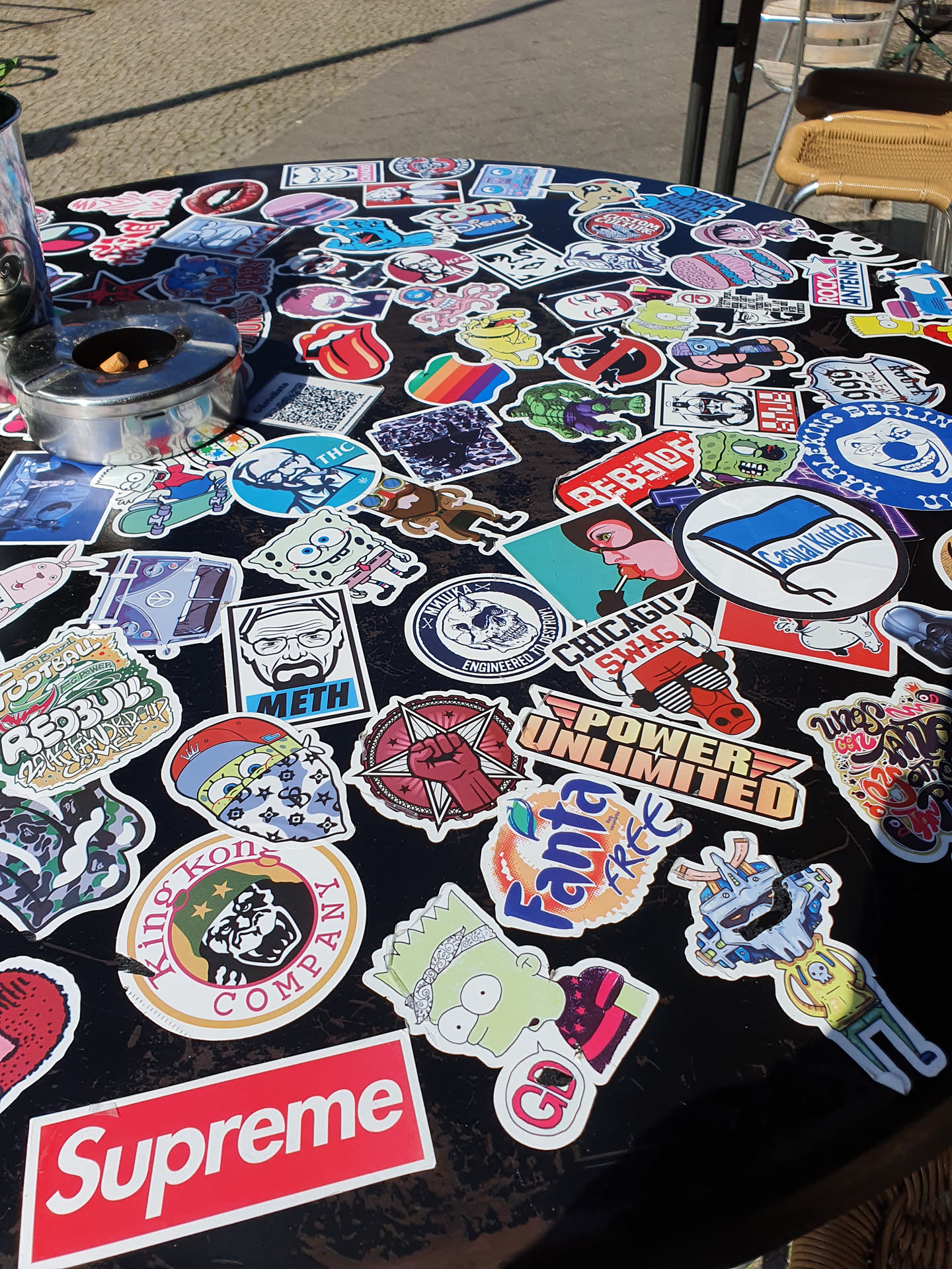 This image shows a table covered in a variety of stickers.  The stickers include various popular brands, cartoon characters, and designs.  A Supreme sticker is prominently featured in the bottom center.