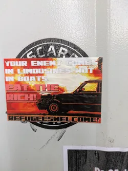 A rectangular sticker with a strong political message. It features the text 'YOUR ENEMY COMES IN LIMOUSINES NOT IN BOATS, EAT THE RICH! REFUGEES WELCOME!' over a background image of a black car against a fiery orange sky.