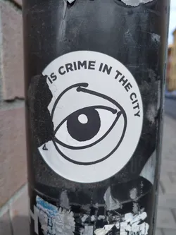 A sticker with a simple illustration of an eye and the text 'Is Crime In The City'. The sticker is partially damaged and is affixed to a dark-colored pole.