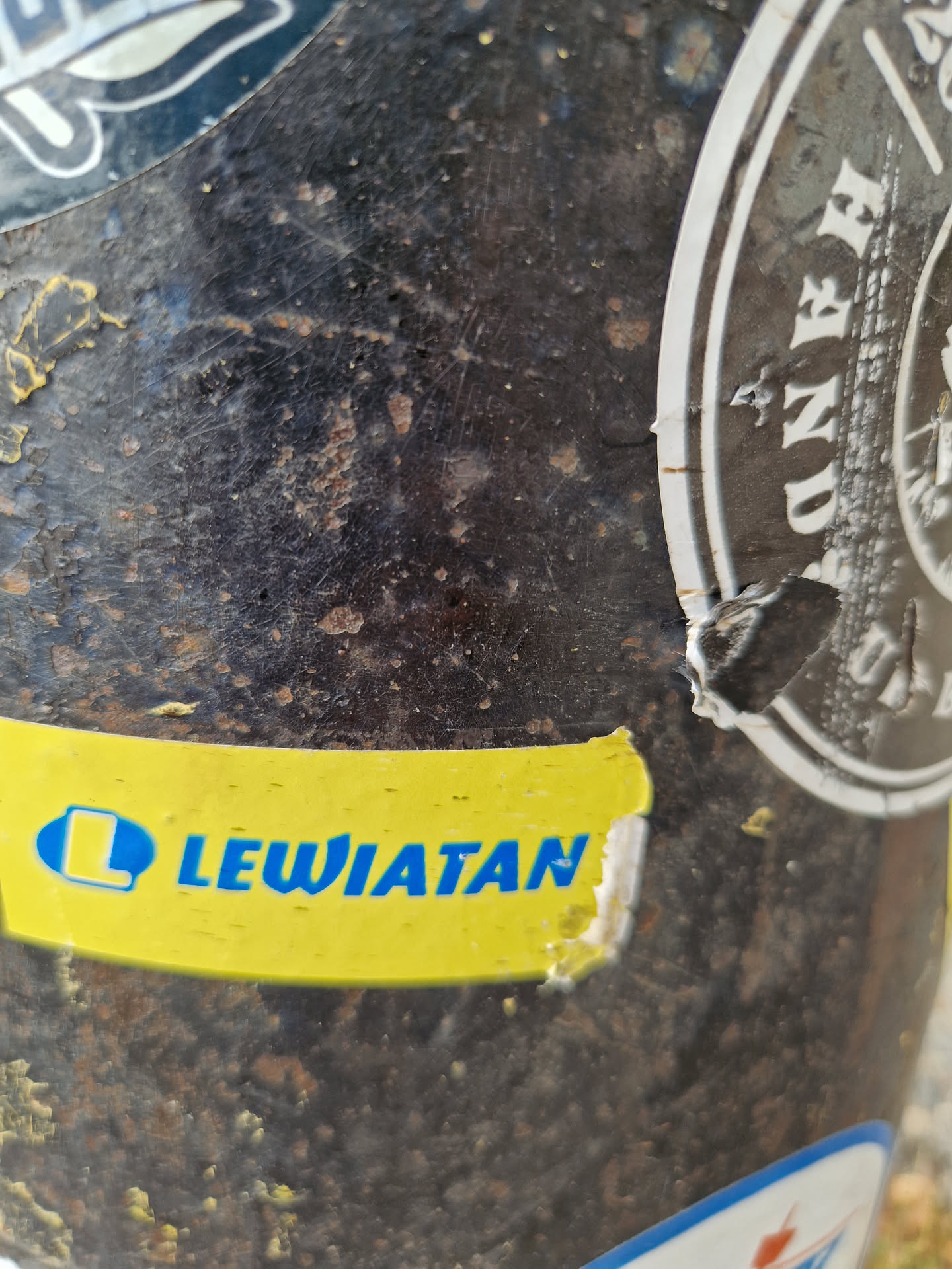A yellow and blue Lewiatan sticker affixed to a dark, textured surface, possibly metal. The sticker shows some wear and tear, indicating exposure to the elements. Other stickers are visible on the same surface.