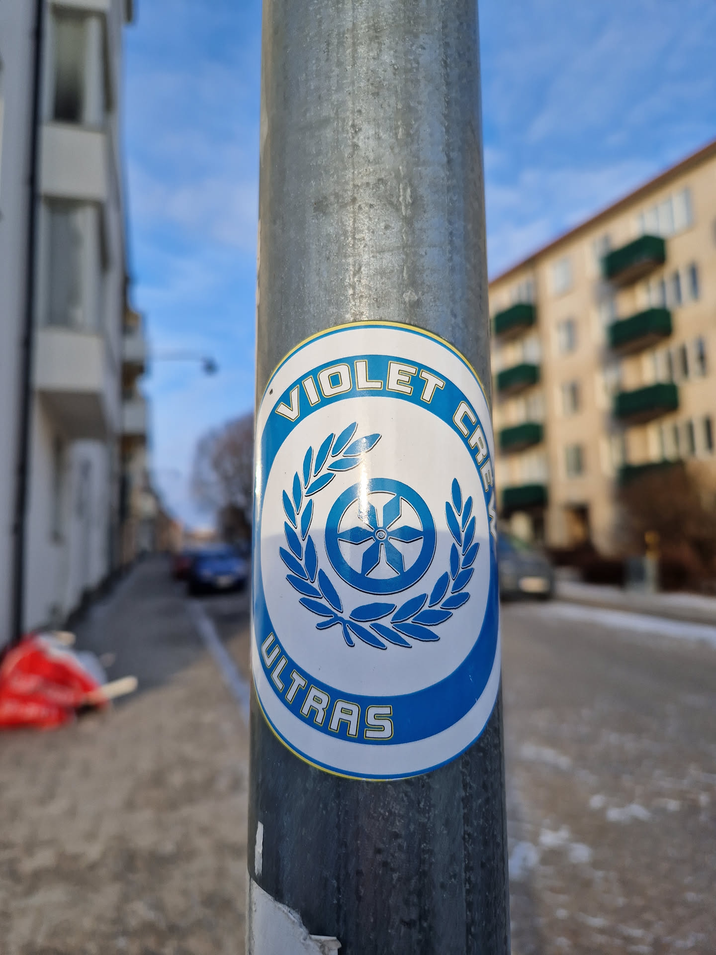 A blue and white circular sticker featuring the text 'Violet Crew Ultras' and a central emblem of a wheel with a flower-like design surrounded by a laurel wreath.  The sticker is affixed to a metal pole.