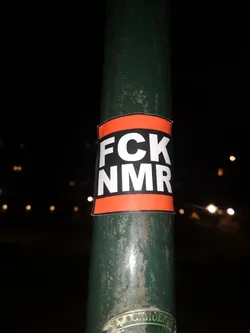 The sticker is rectangular with slightly rounded corners, featuring a prominent black horizontal band in the center. This black band is bordered by two narrower, bright orange horizontal bands at the top and bottom. The text "FCK NMR" is printed in large, bold, white sans-serif capital letters, with "FCK" on the top line and "NMR" on the bottom line, both centered within the black band. The sticker is applied to a dark green, somewhat weathered metal pole, likely a lamppost, at night, with blurred lights in the background.