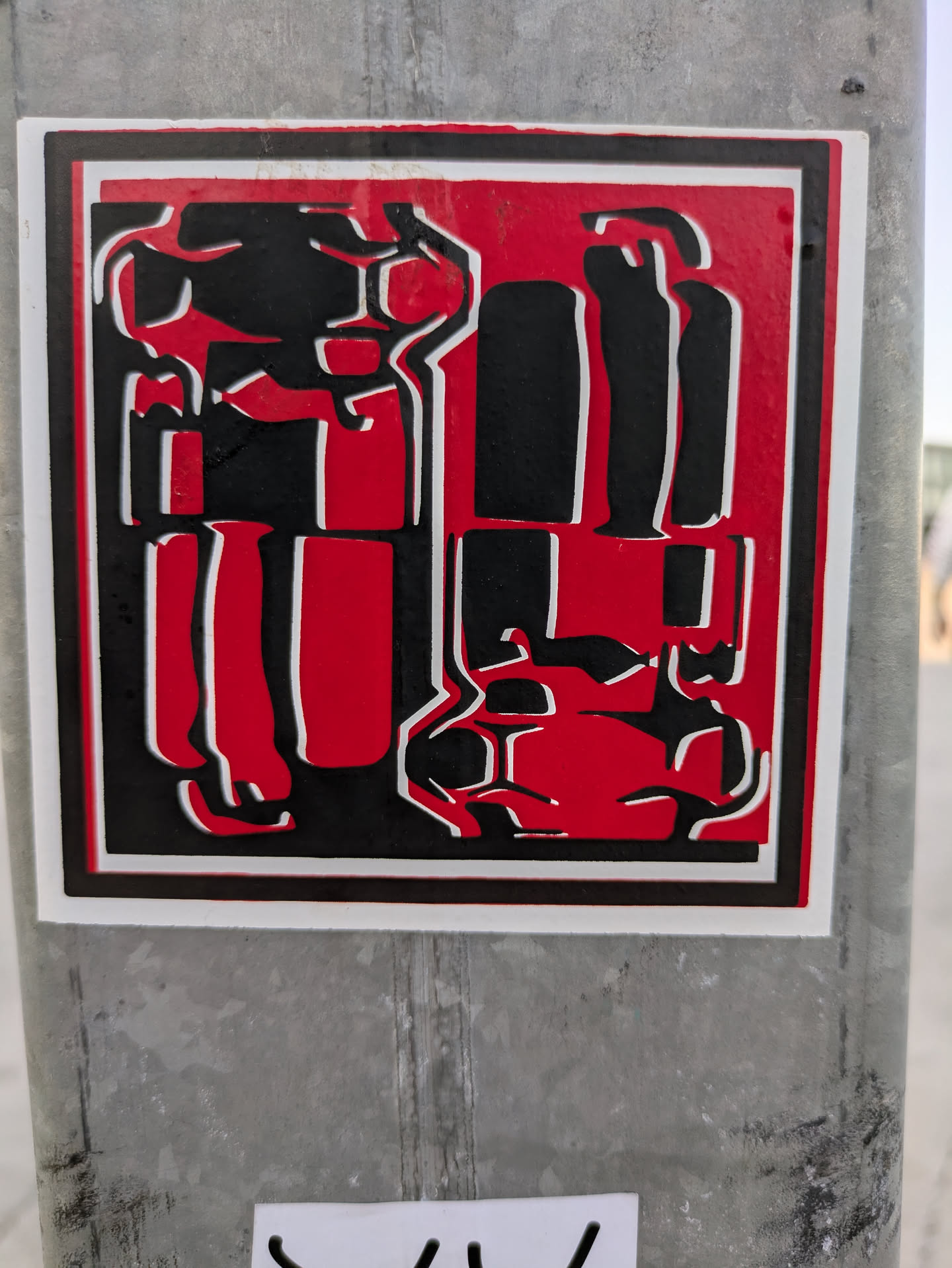 This sticker features an abstract design in red, black, and white. It has a thick black border on a white background. The shapes within the sticker are geometric and somewhat resemble stylized figures or objects. It is affixed to a metal pole.