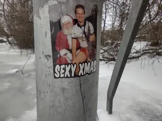 A sticker featuring a man in a Santa outfit and another man with a saxophone sitting on his lap. The sticker has 'SEXY XMAS' written at the bottom.  It's pasted on a metal pole outdoors in a snowy winter setting.