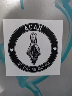 A black and white sticker with a circular design.  The center features an illustration of a clitoris with hands clasped in prayer.  The text "A.C.A.B." is at the top, and "ALL CLITS ARE BEAUTIFUL" at the bottom, within a black circle. Two small hearts are placed on either side of the illustration. 