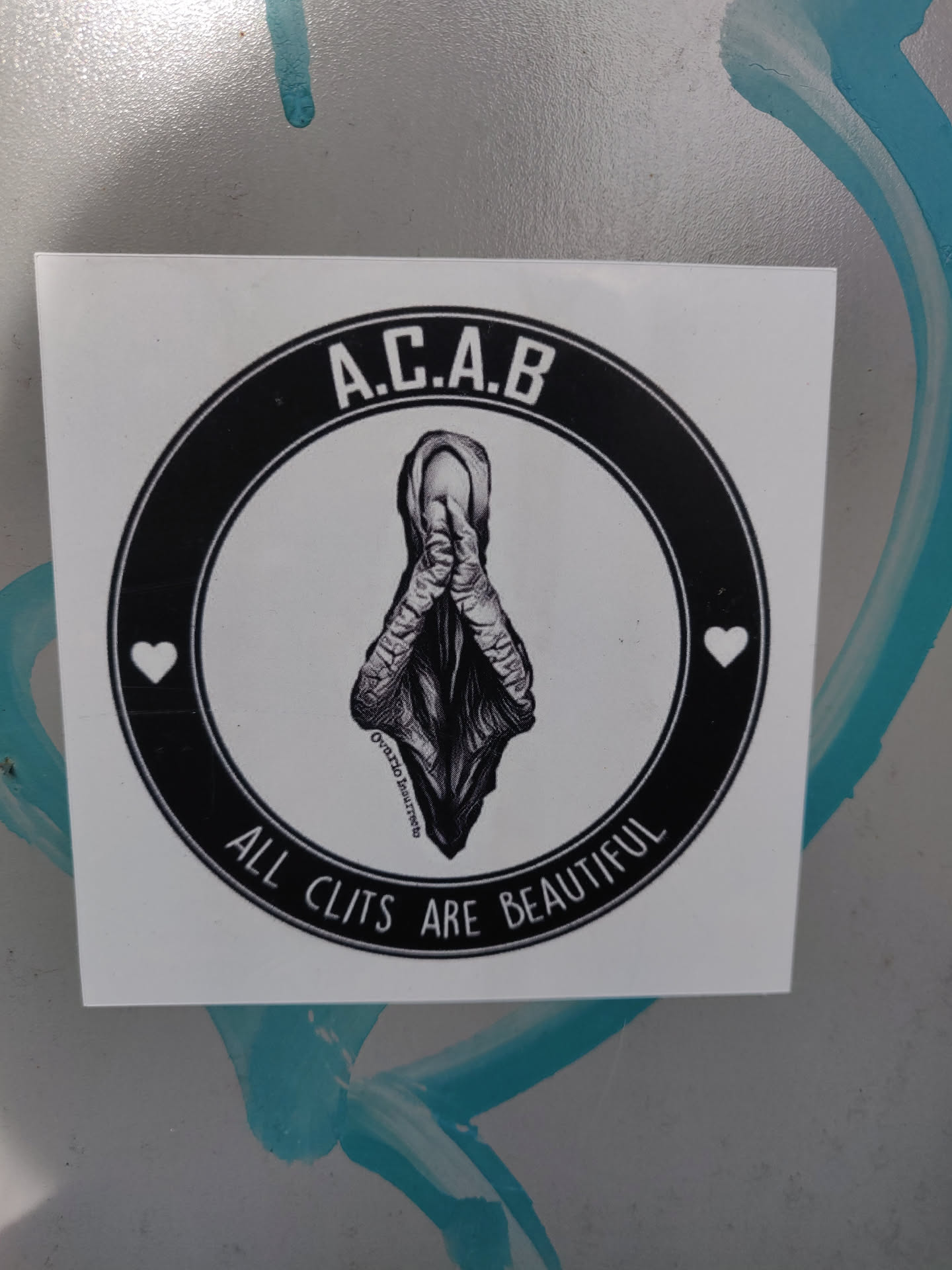 A black and white sticker with a circular design.  The center features an illustration of a clitoris with hands clasped in prayer.  The text "A.C.A.B." is at the top, and "ALL CLITS ARE BEAUTIFUL" at the bottom, within a black circle. Two small hearts are placed on either side of the illustration. 