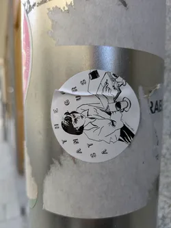 This round sticker features a black and white illustration of a person holding a cup, with a book and other items nearby. The sticker seems to be placed on a metallic pole or surface. The text "A STAN THE FUTURE" is printed around the illustration.