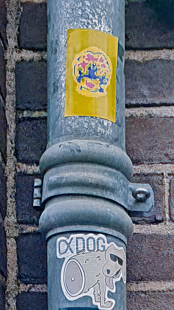 The image features a sticker adhered to a gray metal pole, which is positioned against a brick wall. The sticker has a yellow background with an abstract design in the center, featuring various geometric shapes and colors, including pink, blue, and yellow. Below the sticker is another sticker with a cartoonish dog character and text that reads "CxDog".