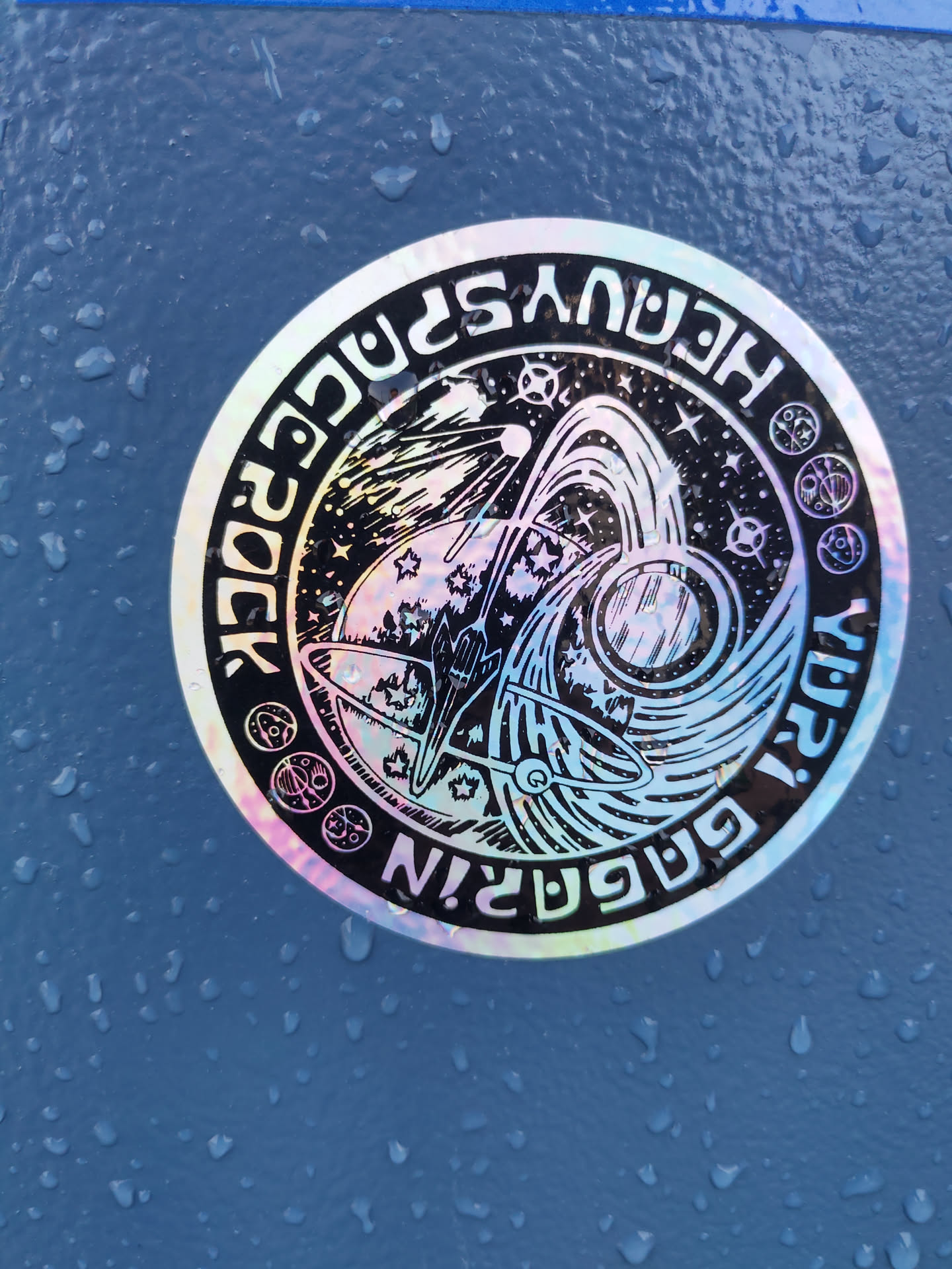 Holographic sticker with a rocket launching into space, surrounded by planets and stars. The text 'Heavy Space Rock!' is written around the circular design.