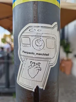 A sticker featuring a drawing of an instant camera printing a picture of a heart. The artist's name Pierpaolo_marchitell is visible on the sticker. 