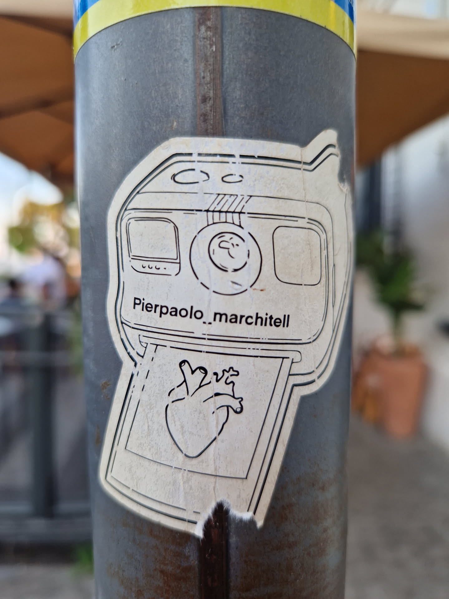 A sticker featuring a drawing of an instant camera printing a picture of a heart. The artist's name Pierpaolo_marchitell is visible on the sticker. 