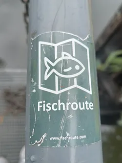A worn green sticker with a fish logo and the text "Fischroute" and a website address. The sticker is affixed to a grey pole outdoors.
